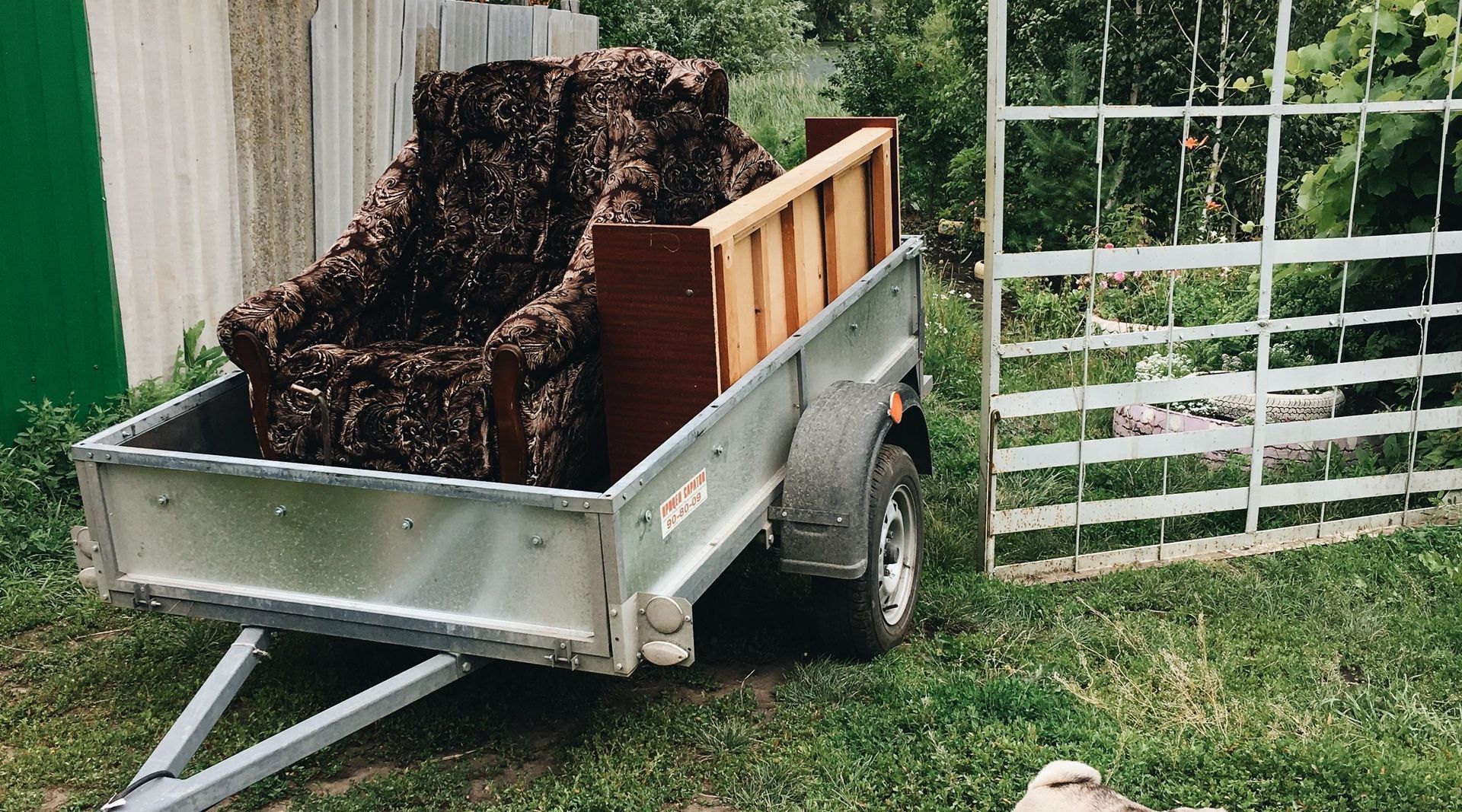 A rural setting with a trailer, vintage armchair, and a pug surrounded by greenery.